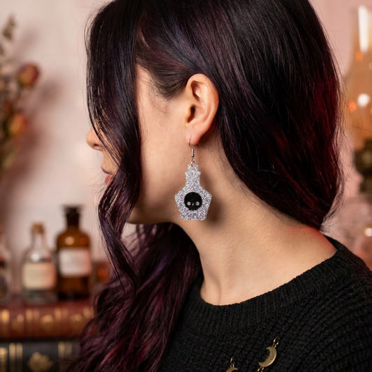 Aretes cortados a laser en forma de pocion magica en acrilico glitter plateado con una calavera grabada usado por modelo en fondo mistico